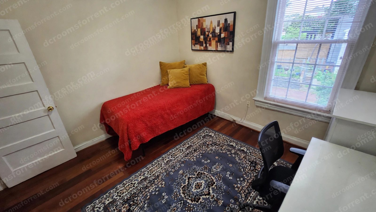Staged Joey Room with red bedspread, mustard pillows, wall art, area rug, and white desk with office chair.