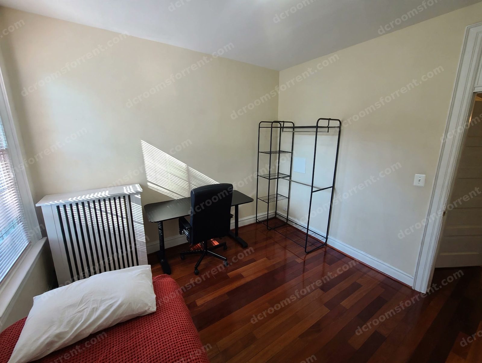 Joey Room with red bedspread and white pillow, radiator, black desk and office chair by the window.