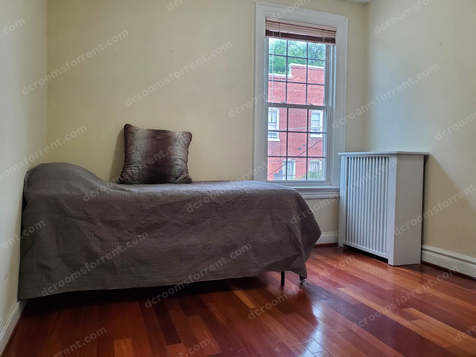 Joey Room with gray coverlet, large window, radiator cover, and hardwood floors.