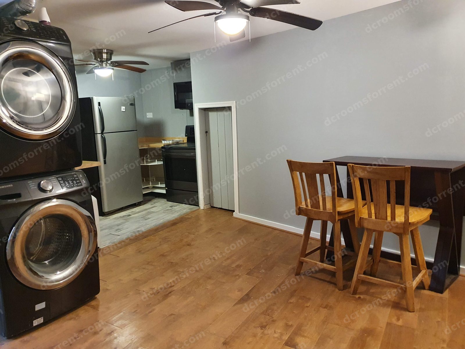 Shared living area with table and stools; kitchen, refrigerator, stove, and stacked washer/dryer in the background.