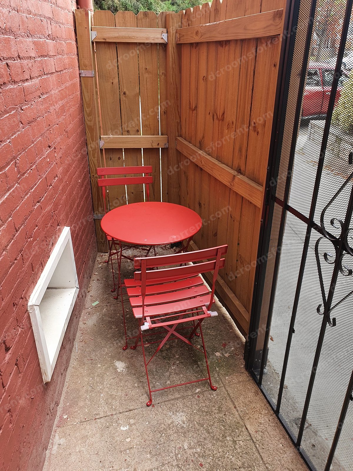Outdoor patio nook with red bistro table and chairs, wooden fence, and security gate.